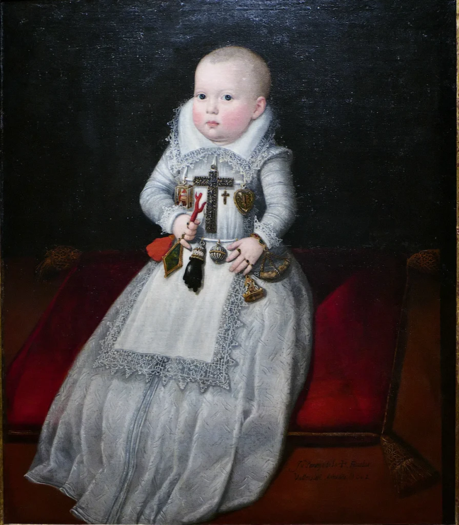 Retrato de una bebé vestida de blanco sobre un cojín rojo, con una gran cruz en el pecho, una rama de coral en la mano y un puño negro de azabache colgado del cinturón